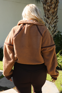 Woman model wearing a tan and brown quarter zip sweater with dark brown athleisure pants. The photo is taken outdoors against some greenery and a white wall. The photo showcases the back of the sweater close up. 