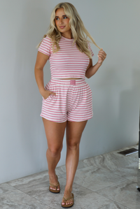 Woman wearing a pink and multi-colored striped patterned two piece set featuring a cropped t-shirt and high waisted, elastic waist shorts indoors against a white wall with white marble flooring. 