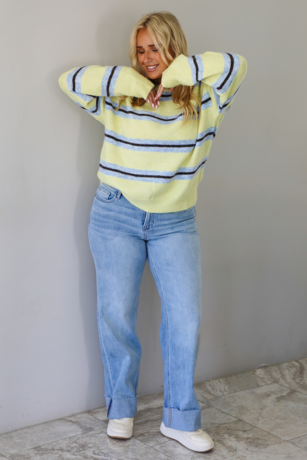 Woman wearing a pastel yellow, blue, and dark brown striped sweater, paired with light wash denim jeans that have a wide leg cut with a cuffed hem, standing indoors against a white wall with white marble flooring.