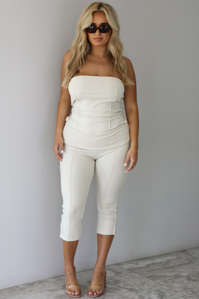 Woman wearing a cream two piece set featuring a strapless tube top and capri pants that fold over at the waist indoors against a white wall with white marble flooring. 