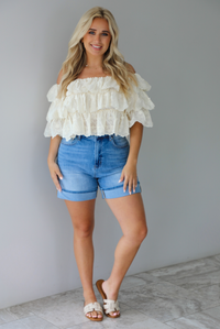 Woman wearing a cream off the shoulder, ruffled eyelet cropped top with medium wash denim shorts indoors against a white wall with white marble flooring. 