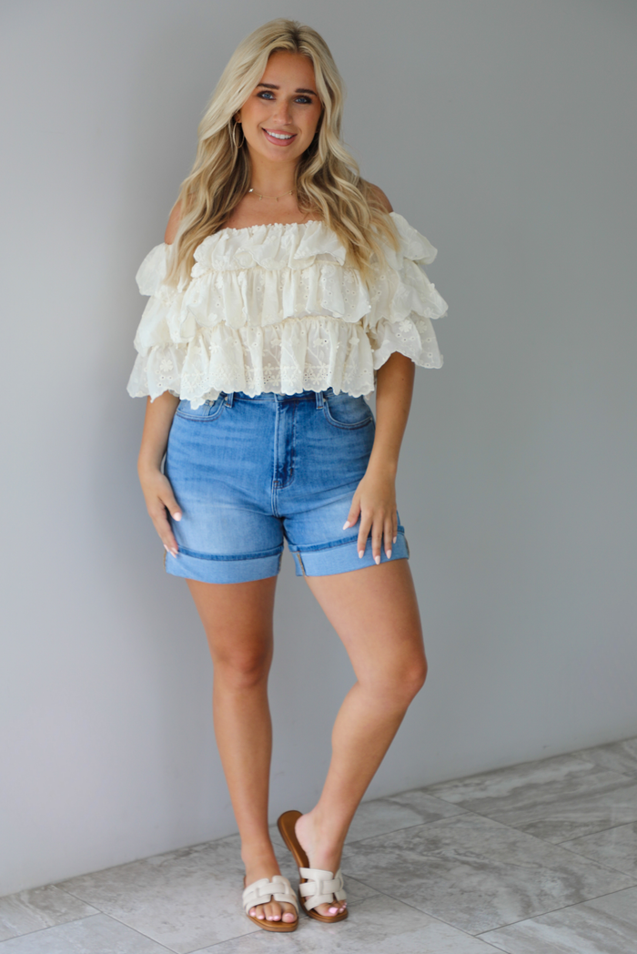 Woman wearing a cream off the shoulder, ruffled eyelet cropped top with medium wash denim shorts indoors against a white wall with white marble flooring. 
