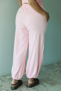 Woman wearing a pink two piece set featuring a cropped short sleeved top and high waisted jogger-style pants indoors against a white wall with white marble flooring. The photo shows a closeup of the back of the pants.