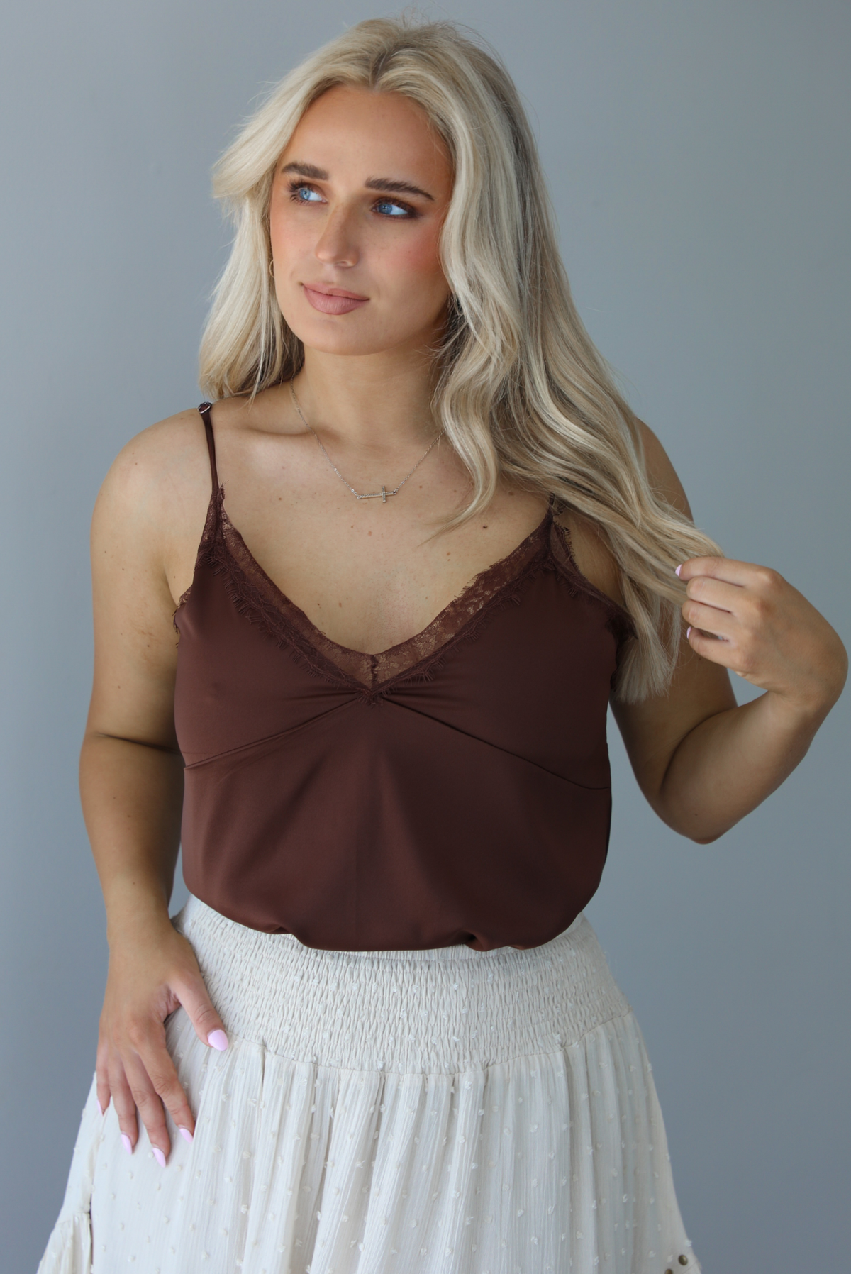 Woman wearing a satin brown tank that is tucked into a high waisted sand colored midi length skirt. The photo is taken in front of a white wall.