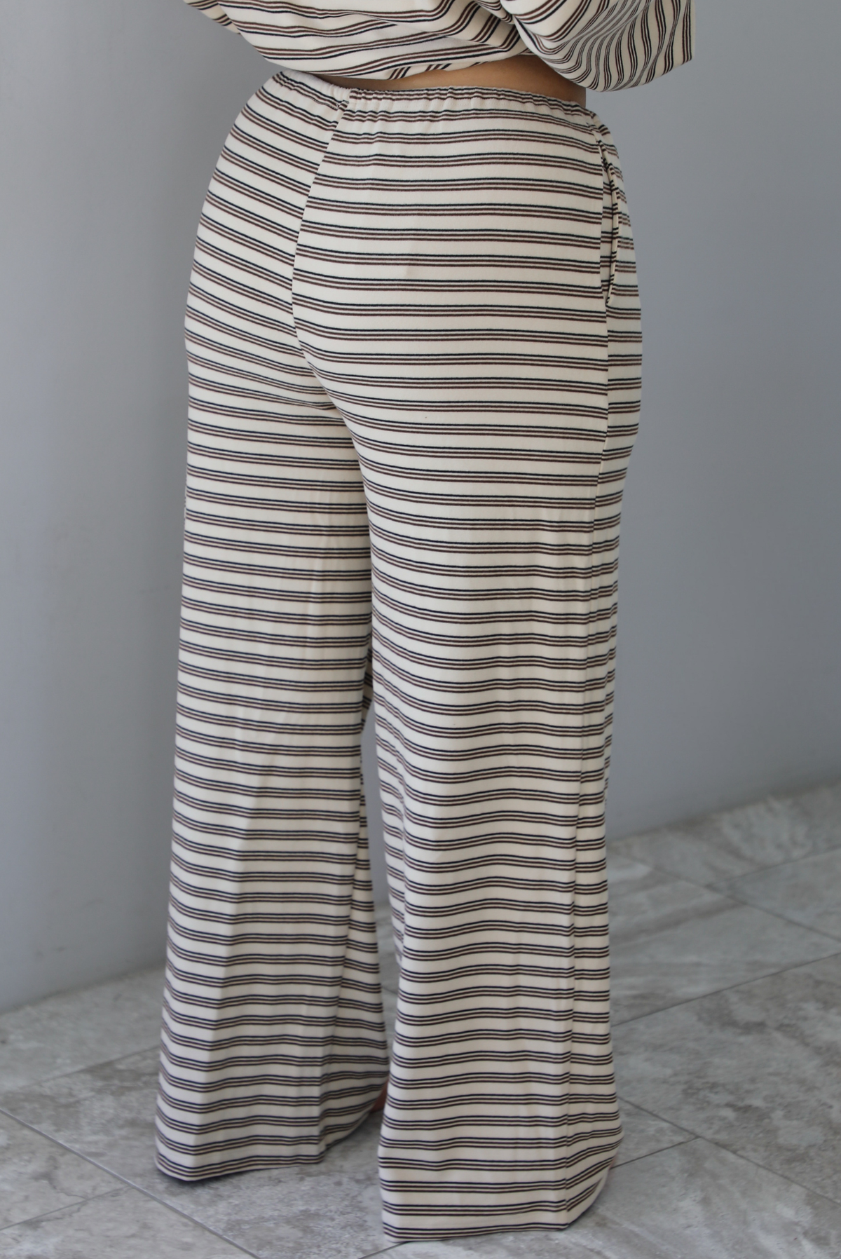 A woman shown from the waist down from behind wearing a sand colored matching two piece set with a brown striped pattern throughout featuring an oversized long sleeved top and oversized slouchy wide leg pants, standing indoors against a white wall with white marble flooring. The photo focuses on the back of the pants.