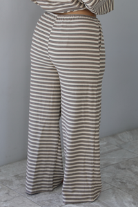 A woman shown from the waist down from behind wearing a sand colored matching two piece set with a brown striped pattern throughout featuring an oversized long sleeved top and oversized slouchy wide leg pants, standing indoors against a white wall with white marble flooring. The photo focuses on the back of the pants.