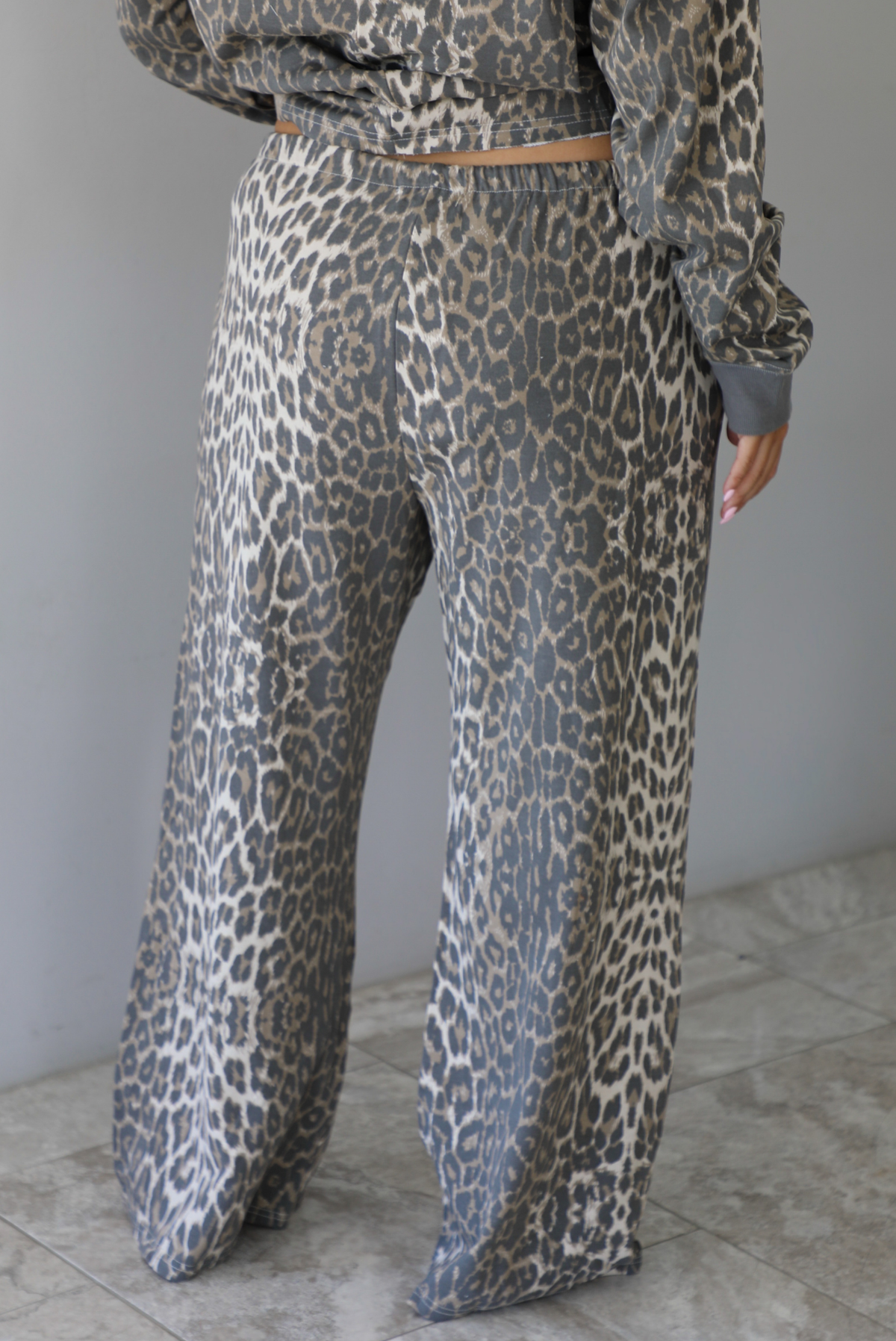 A woman wearing a leopard patterned two piece set featuring an oversized top with a front pocket detail and mid-rise oversized pants, standing indoors against a white wall with white marble flooring. The photo shows a closeup of the back of the pants. 
