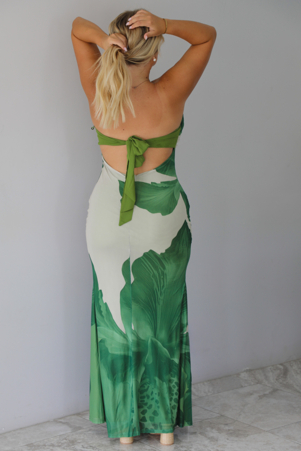 A blonde woman shown from behind models a strapless, bodycon maxi dress with a green tropical leaf print on a light beige background, with a tie closure in the back, standing indoors against a white wall with white marble flooring. 