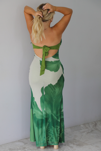 A blonde woman shown from behind models a strapless, bodycon maxi dress with a green tropical leaf print on a light beige background, with a tie closure in the back, standing indoors against a white wall with white marble flooring. 