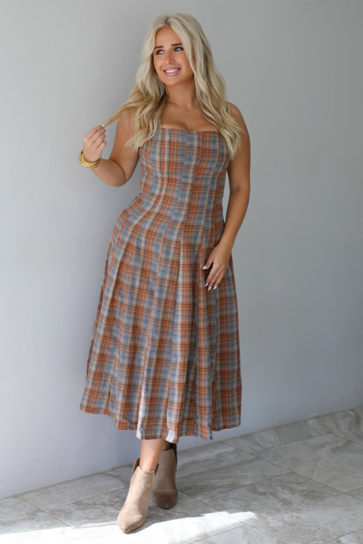 A woman wearing a brown and blue plaid midi dress with thick straps, a square neckline, fitted bodice that starts flaring out at the hips and ends about mid-shin, with a flowy skirt, standing indoors against a white wall with white marble flooring. 