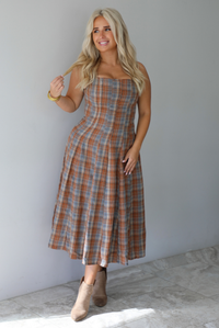 A woman wearing a brown and blue plaid midi dress with thick straps, a square neckline, fitted bodice that starts flaring out at the hips and ends about mid-shin, with a flowy skirt, standing indoors against a white wall with white marble flooring. 
