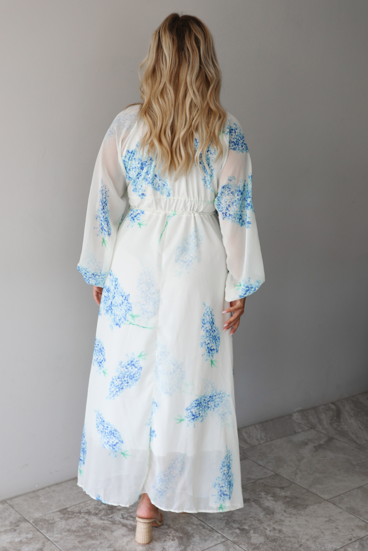 A woman shown from behind modeling a white maxi dress with a blue floral print and a sheer, flowing overlay, focusing on the bodice of the dress with its cinched waistline, standing indoors against a white wall with white marble flooring. 
