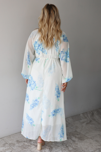A woman shown from behind modeling a white maxi dress with a blue floral print and a sheer, flowing overlay, focusing on the bodice of the dress with its cinched waistline, standing indoors against a white wall with white marble flooring. 