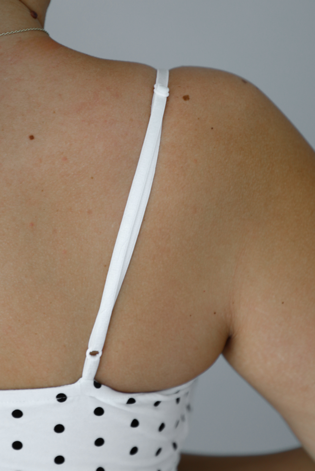 Woman wearing a white dress with a black polka dot print pattern throughout, spaghetti straps, a fitted design, mini dress cut, with a ruffled hem indoors against a white wall. The photo shows a closeup of the adjustable straps in the back. 