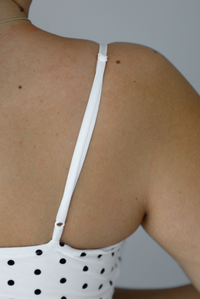 Woman wearing a white dress with a black polka dot print pattern throughout, spaghetti straps, a fitted design, mini dress cut, with a ruffled hem indoors against a white wall. The photo shows a closeup of the adjustable straps in the back. 
