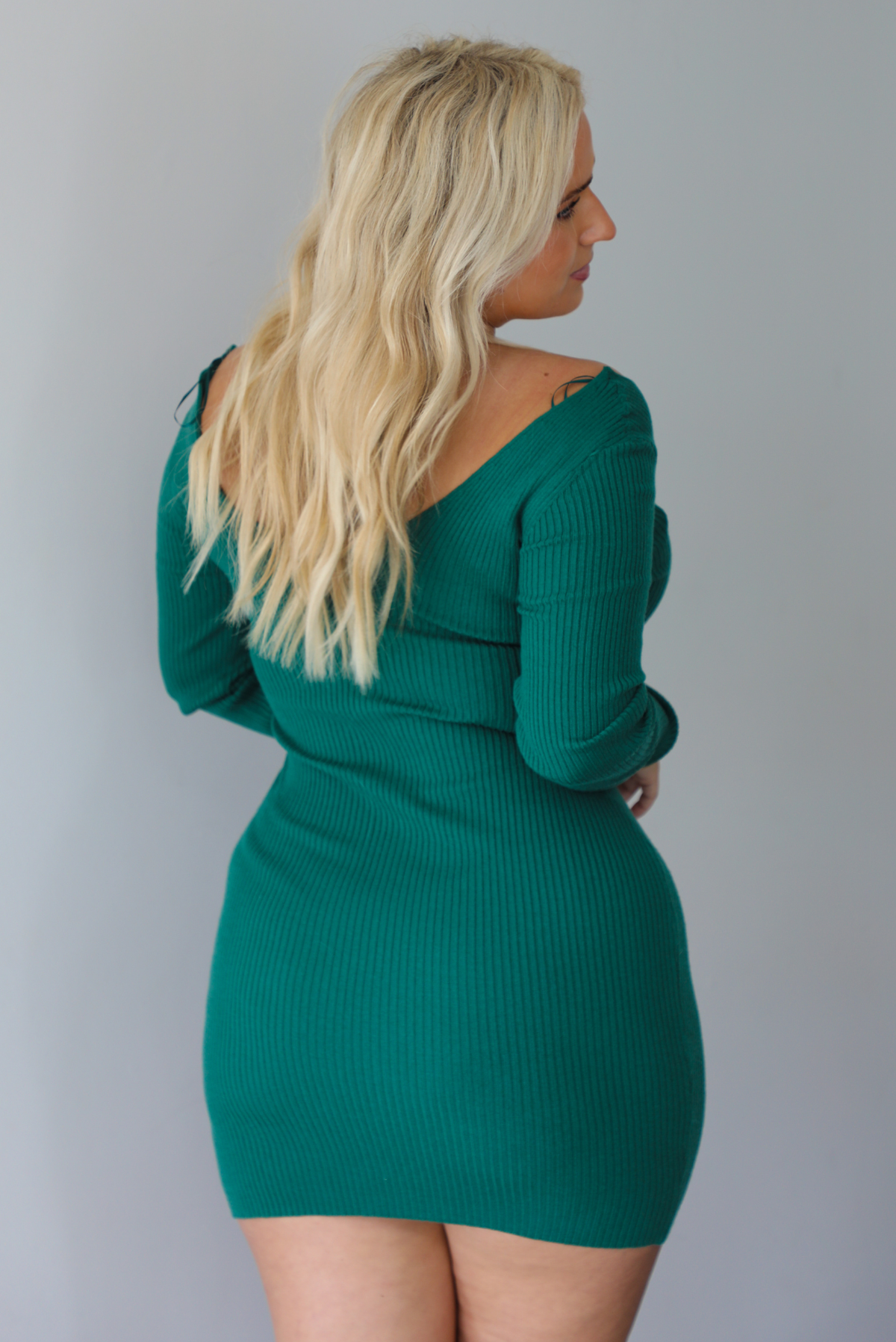 A woman shown from behind wearing a hunter green mini dress that has long sleeves, a v-neckline, a ribbed texture to the material, and is fitted, standing indoors against a white wall.