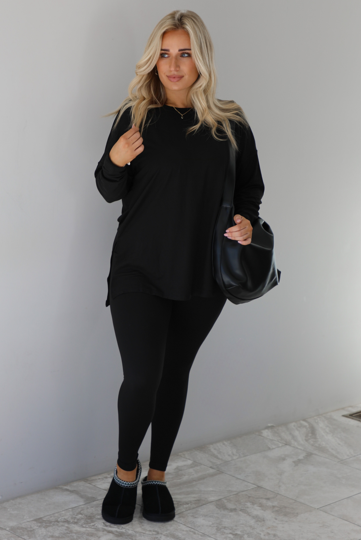 Woman wearing a black two piece set that features an oversized long sleeved top and fitted leggings. The set is paired with a black vegan leather shoulder bag and black slide on slippers. The photo is taken indoors against a white wall with white marble flooring.