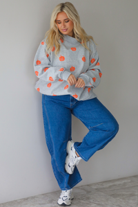 A woman wearing a heather grey oversized sweatshirt that has a repeating jack-o-lantern design throughout, paired with dark wash denim jeans, standing indoors against a white wall with white marble flooring. 