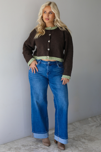 A woman wearing a dark brown with green trim cardigan fully buttoned up paired with dark wash, cuffed hem denim jeans, standing indoors against a white wall with white marble flooring. 