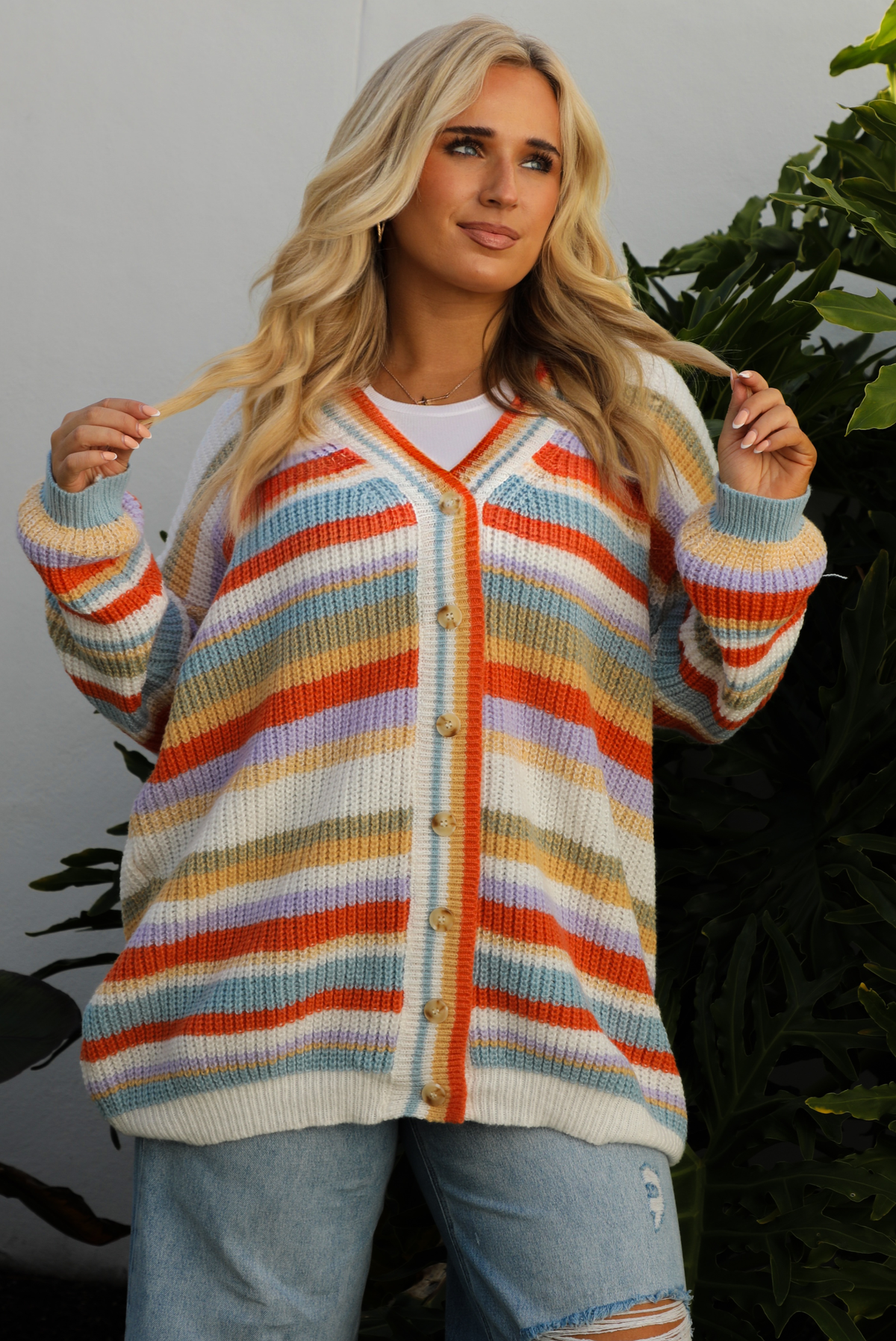 Woman model wearing an orange, lavender, yellow, muted green, blue, and white striped oversized cardigan with functional button closures in the front and an oversized fit with a light wash flared hem denim. The photo is taken outdoors against a white wall and greenery in the back. The photo showcases a close up of the cardigan and shows the full length, which falls to the upper thigh on our 5'7" model.