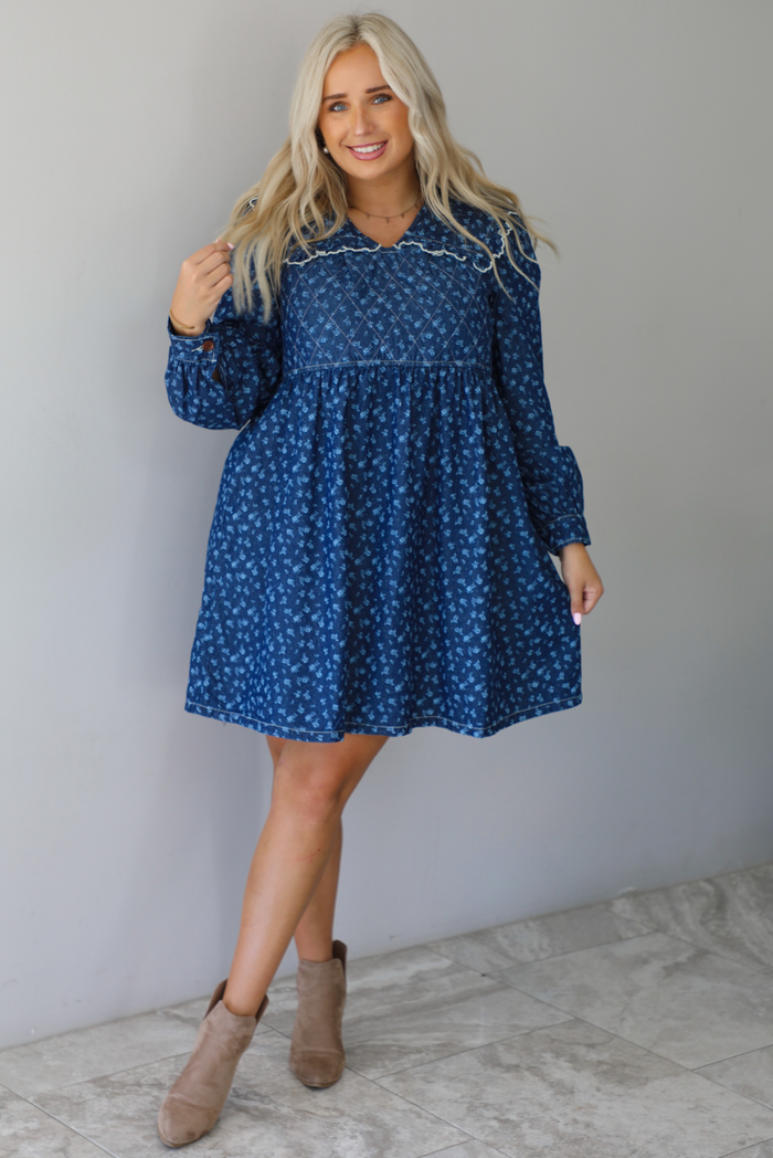 A woman wearing a dark denim mini dress with a white floral print pattern throughout, collared neckline, a quilted detail at the bodice, long sleeves, and a flowy skirt, standing indoors against a white wall with white marble flooring. 