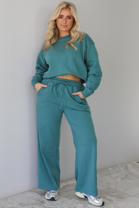 Woman wearing a sage two piece sweat-set. The set features a slightly oversized sweater and high waisted sweat pants that have an elastic and drawstring waistline with pockets. The photo is taken indoors against a white wall with white marble flooring. 