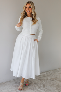 Woman wearing a white two piece set featuring a long sleeved cardigan and a high waisted midi length skirt. The photo is taken indoors against a white wall with white marble flooring. 