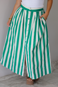 Closeup of a woman wearing a white tank top and a maxi length cream and green striped skirt, focused in on the skirt, which has a high waistline, functional pockets on either side, functional button closures in the front, and has a flowy fit, standing indoors against a white wall with white marble flooring. 