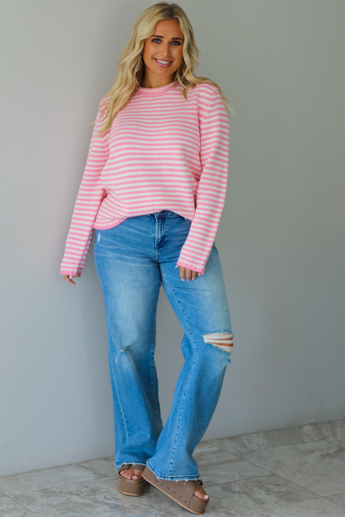 A woman wearing a pink and white striped sweater paired with light wash denim jeans, standing indoors against a white wall with white marble flooring. 