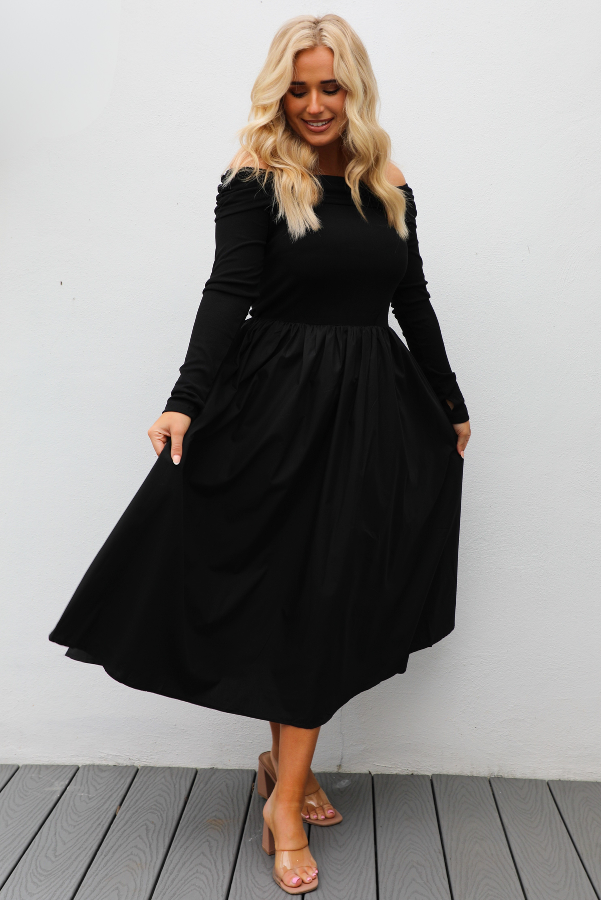 Woman wearing a black off the shoulder, long sleeved midi length dress with a pair of clear strapped nude heels. The photo is taken outdoors against a white wall.