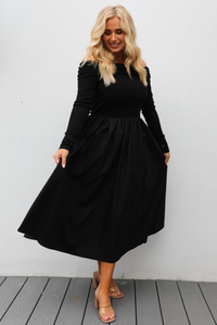 Woman wearing a black off the shoulder, long sleeved midi length dress with a pair of clear strapped nude heels. The photo is taken outdoors against a white wall.