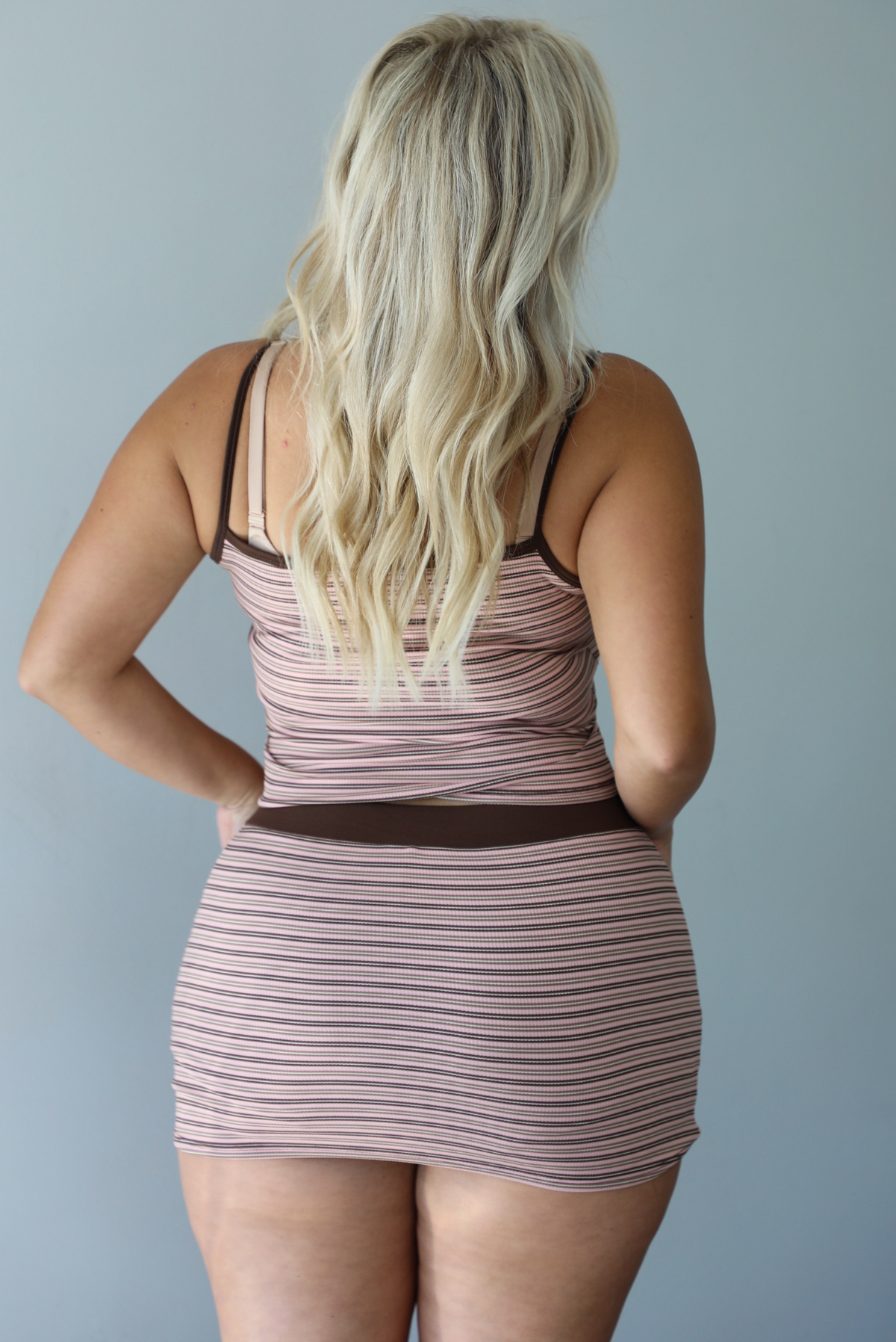 A woman shown from behind wearing a pink and brown striped two piece matching set consisting of a tank top and mini skirt with a thick brown waistband, standing indoors against a white wall.