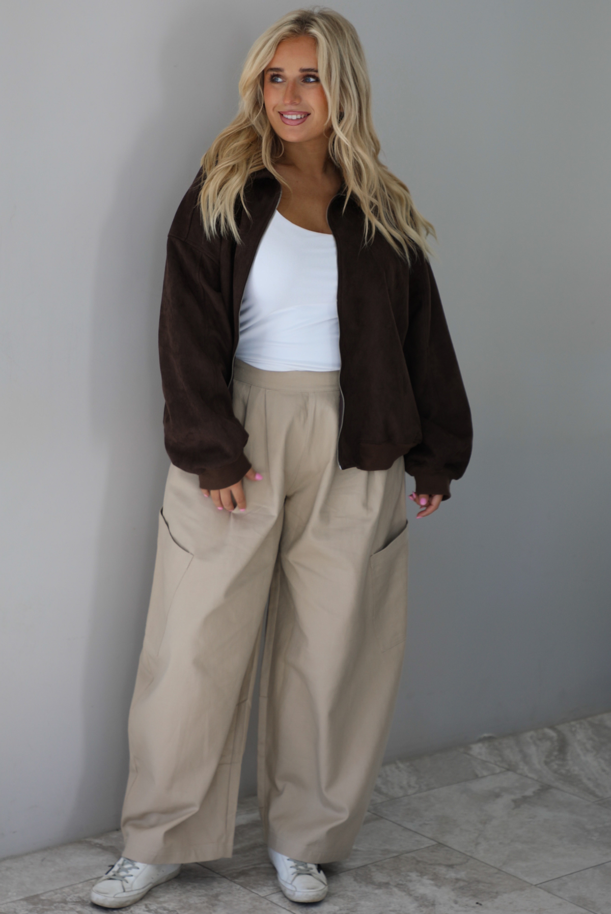 A woman wearing a brown suede jacket with a white tank and sand colored baggy pants, standing indoors against a white wall with white marble flooring. 