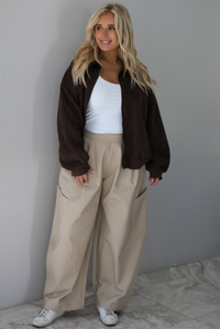 A woman wearing a brown suede jacket with a white tank and sand colored baggy pants, standing indoors against a white wall with white marble flooring. 