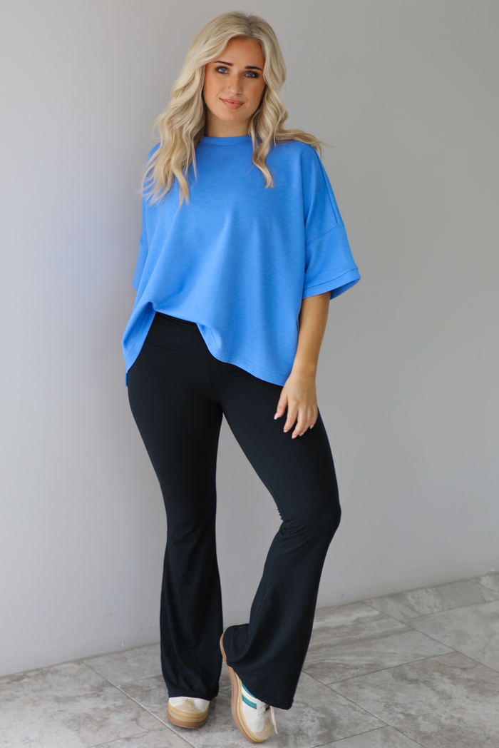 Woman wearing an oversized blue box tee with black leggings that flare slightly at the hem. The photo is taken indoors against a white wall with white marble flooring. 