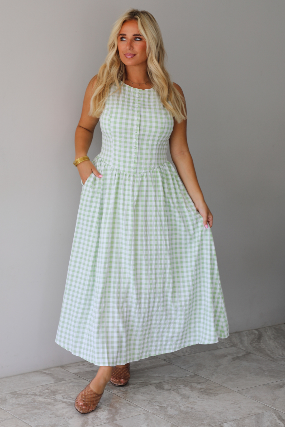 A woman wearing a light green and white gingham maxi dress with a drop waist bodice and side pockets, standing indoors against a white wall with white marble flooring. 