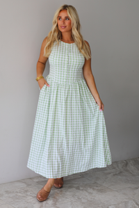 A woman wearing a light green and white gingham maxi dress with a drop waist bodice and side pockets, standing indoors against a white wall with white marble flooring. 