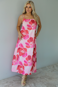 Woman wearing a pink and red floral print maxi dress with thin straps and a flowy fit indoors against a white wall with white marble flooring. 