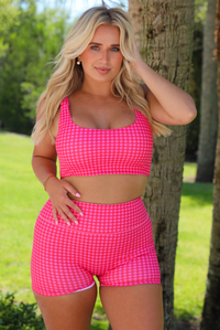 A woman wearing a red and hot pink gingham two-piece activewear set consisting of a crop top and matching shorts, outdoors with greenery and palm trees in the background. 