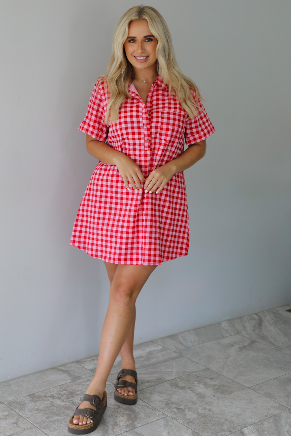 Woman wearing a pink and red gingham collared neckline t-shirt dress indoors against a white wall with white marble flooring. 