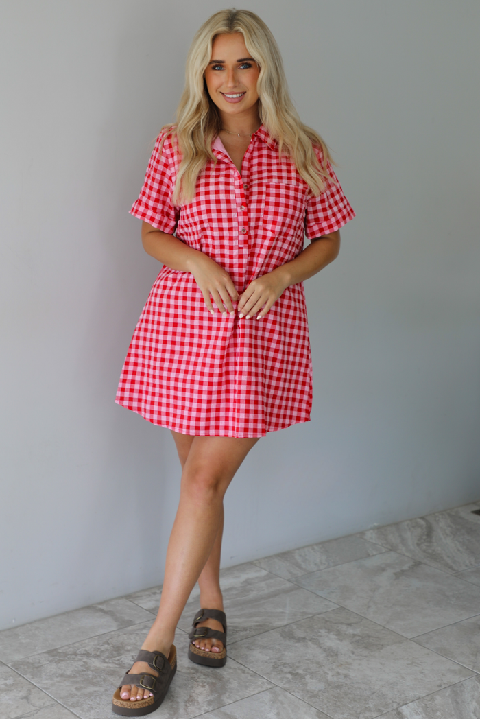 Woman wearing a pink and red gingham collared neckline t-shirt dress indoors against a white wall with white marble flooring. 
