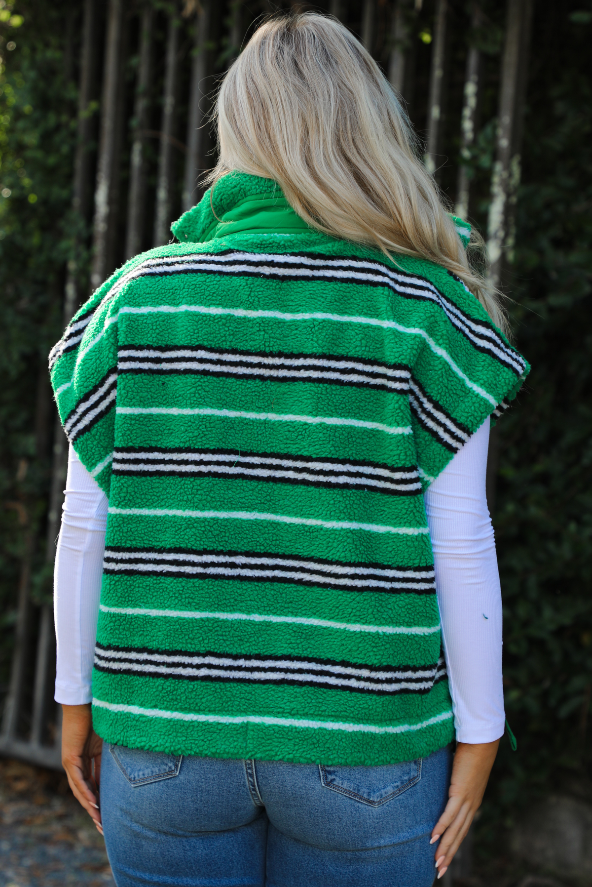 Woman model wearing a green, black, and white striped vest that has a sherpa texture to it, functional button closures in the front, and tie closures on either side, It is paired with a white long sleeved top, a medium wash pair of denim jeans, and tan ugg boots. The photo is taken outdoors with a metal gate and greenery in the back. The back if the vest is shown, displaying the high collar in the back and the length of the vest, which stops mid-hip on the 5'7" model.