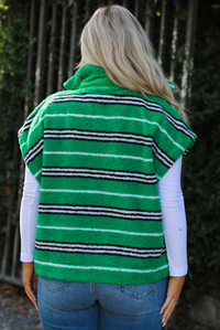 Woman model wearing a green, black, and white striped vest that has a sherpa texture to it, functional button closures in the front, and tie closures on either side, It is paired with a white long sleeved top, a medium wash pair of denim jeans, and tan ugg boots. The photo is taken outdoors with a metal gate and greenery in the back. The back if the vest is shown, displaying the high collar in the back and the length of the vest, which stops mid-hip on the 5'7" model.
