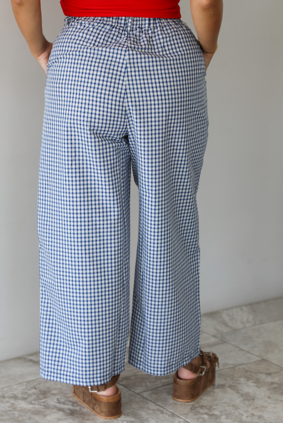 Woman wearing a navy and white plaid print pants that have a high waist, pleated details, functional pockets on either side, and a professional fit indoors against a white wall with white marble flooring. The photo shows the back of the pants. 