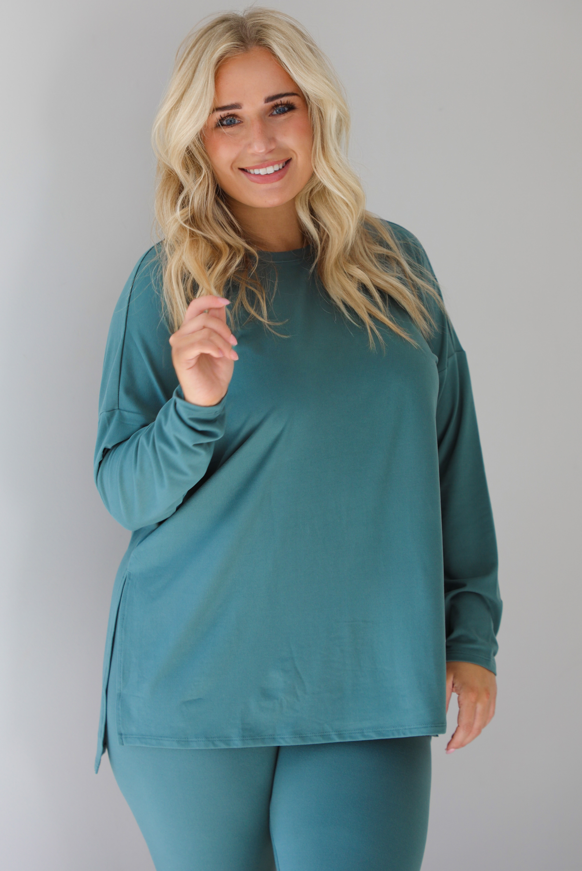 Woman wearing a teal two piece set that features an oversized long sleeved top and fitted leggings. The photo shows a closeup of the top and shows how it has split hem details on either side. The photo is taken indoors against a white wall.