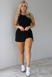 Woman wearing a black athleisure romper that has a wrap-style skirt detail that wraps around and ties in the front on the left side. It's styled with white sneakers that have black stripes down the side. The photo is taken indoors against a white wall with white marbled flooring. 