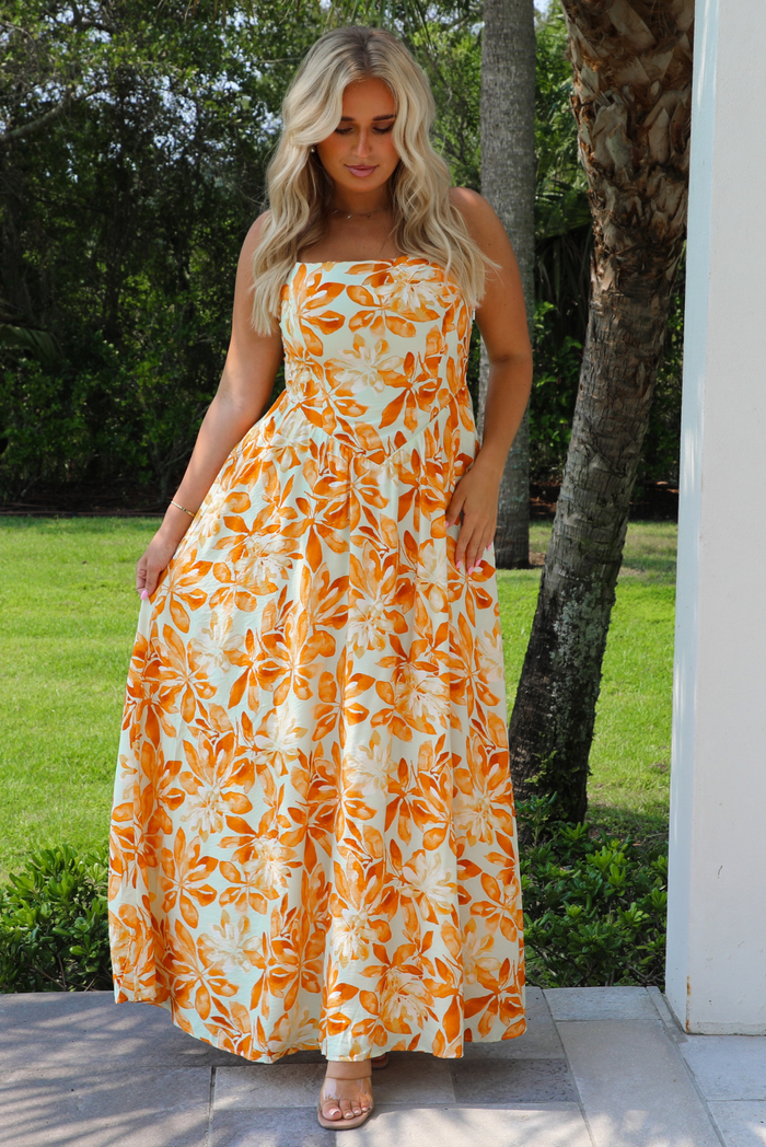 A woman wearing a spaghetti strapped maxi dress with a vibrant marigold orange and yellow floral pattern on a white background, standing outdoors on a patio with green grass and trees in the background.