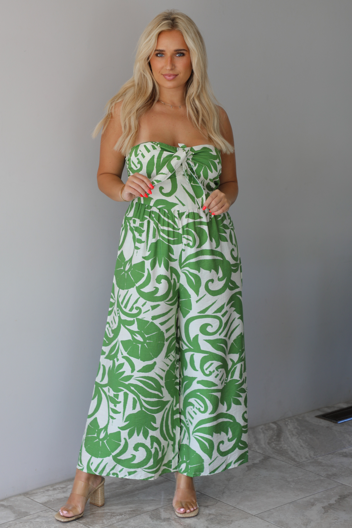 Woman wearing a strapless jumpsuit with green leaf pattern indoors against a white wall with white marble flooring. 
