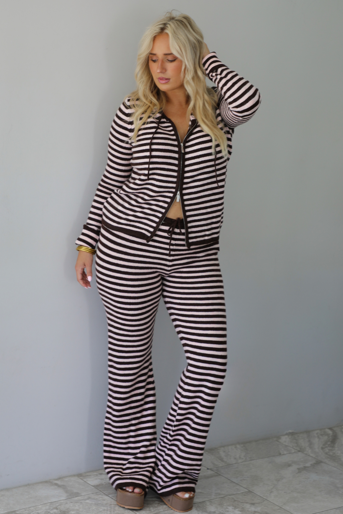 A woman wearing a light pink and brown striped two piece set featuring a double zipped hoodie top and mid-rise, wide leg pants, standing indoors against a white wall with white marble flooring. 