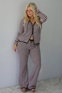 A woman wearing a light pink and brown striped two piece set featuring a double zipped hoodie top and mid-rise, wide leg pants, standing indoors against a white wall with white marble flooring. 